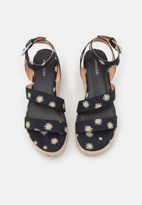 Een paar zwarte sandalen met witte en gele madeliefjespatronen, enkelbandjes met gespen en gevlochten wiggen op een witte achtergrond.