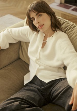 Cardigan bianco in maglia con bottoni sul davanti, caratterizzato da una vestibilità ampia. Abbinato a pantaloni scuri a righe sottili, seduto su un divano beige con texture.