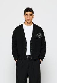 Cardigã preto de malha com decote em V, mangas longas e fecho de botões. Apresenta um logotipo cinza do lado esquerdo. Usado sobre uma camisa branca.