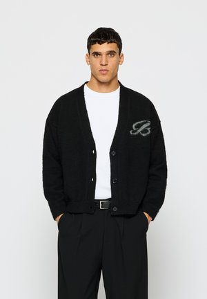 Cardigan en tricot noir avec un col en V, des manches longues et une fermeture à boutons. Présente un logo gris sur le côté gauche. Porté sur une chemise blanche.