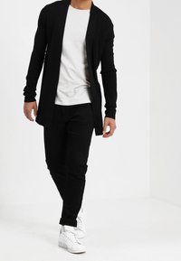 Svart lång cardigan, mjukt stickad tyg, öppen framsidan. Bärs över en vit t-shirt och svarta byxor, kombinerat med vita sneakers.