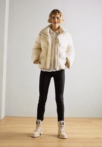 Giacca trapuntata beige con colletto alto e chiusura a zip, indossata sopra una camicia bianca, abbinata a pantaloni neri aderenti e stivaletti color crema.