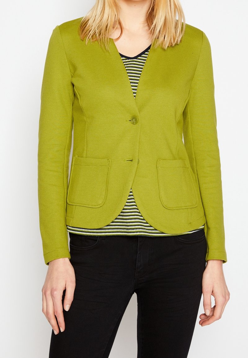 Blazer vert texturé avec un seul bouton, deux poches avant et une coupe ajustée, superposé à un haut rayé, associé à un pantalon noir.