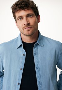 Světle modrá džínová košile s knoflíkovým zapínáním, límcem a jemnou texturou, nošená přes černé tričko. Žádné vzory ani akcenty nejsou viditelné.