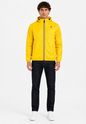 Homme debout, droit, portant une veste à capuche jaune vif, un jean foncé et des baskets blanches sur un fond blanc uni.