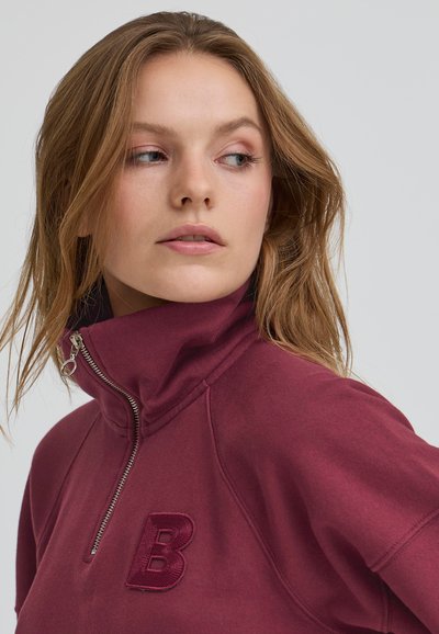 Burgundska zip-up trenirka z visokim ovratnikom, ki vsebuje vezeno črko "B" in srebrno zadrgo, izdelana iz gladkega, teksturiranega materiala.