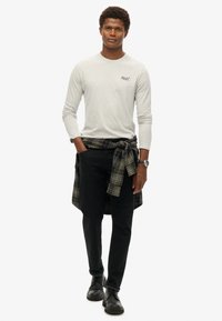 Grijze longsleeve shirt met een logo, zwarte jeans en zwarte schoenen. Een groen en blauw geruit shirt om de taille gebonden voegt textuur toe.