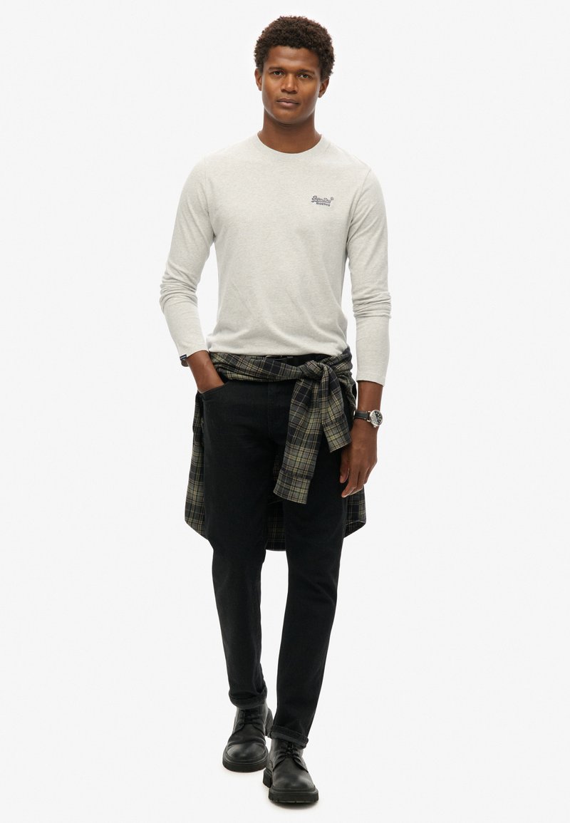 Grijze longsleeve shirt met een logo, zwarte jeans en zwarte schoenen. Een groen en blauw geruit shirt om de taille gebonden voegt textuur toe.