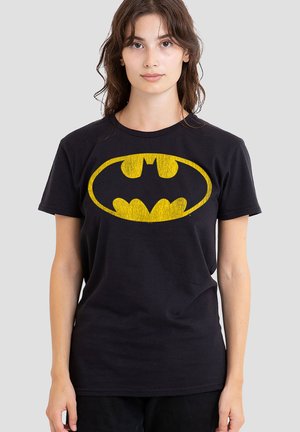 Young woman wearing a black t-shirt with a large yellow Batman logo on the front, standing against a plain gray background.