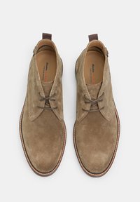 Zapatos de ante marrón con punta redondeada, que cuentan con un diseño de cordones, un pequeño lazo marrón en el talón y una suela de puntadas en contraste.