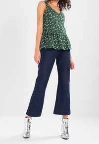 Top verde con peplum y patrón floral blanco, combinado con vaqueros acampanados azul oscuro y botines plateados metálicos.