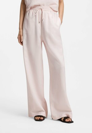 Personne portant un pantalon large rose clair avec une taille élastique et un cordon, associé à des sandales noires à bout ouvert, sur fond blanc.