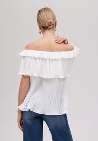 Weißes, plissiertes Top mit schulterfreiem Design und Rüschensaum, das eine lockere Passform und eine weiche Textur bietet. Kombiniert mit blauen Denim-Jeans.