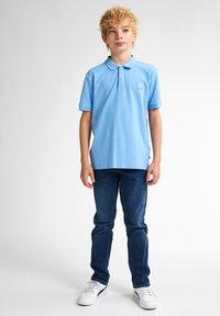 Světle modré polo tričko s límečkem a malou nášivkou, v kombinaci s tmavě modrými džíny a bílými teniskami. Jednoduchý, pohodlný design.