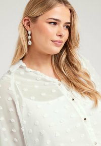 Blusón blanco transparente con un patrón de lunares texturizados, que presenta un cierre frontal de botones y un cuello curvado. Cabello largo y ondulado de color rubio y pendientes llamativos.