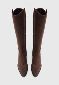 Botas de ante marrón por encima de la rodilla con punta afilada y cremallera lateral. Presentan una textura suave y una suela negra contrastante.