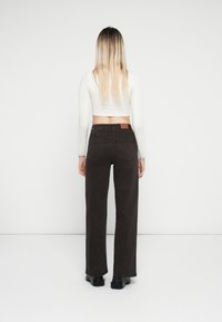 Bruine hoge taille wijde broek met achterzakken, gecombineerd met een witte lange mouwen crop top, tegen een witte achtergrond.