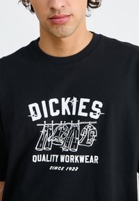 Juodas medvilninis marškinėliai su baltu grafiniu dizainu, vaizduojančiu kabantį darbo rūbą, su tekstu "Dickies Quality Workwear" ir "Since 1922."
