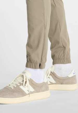Beige sneakers with white accents and cream laces. Textured suede upper. Light beige elastic cuffs on pants, featuring zip detail at hem.