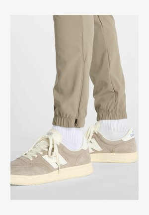 Beige Sneaker mit weißen Akzenten und cremefarbenen Schnürsenkeln. Obermaterial aus strukturiertem Wildleder. Helle beige elastische Bündchen an der Hose, mit Reißverschlussdetails am Saum.