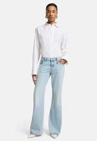 Chemise blanche à manches longues avec un col pointu, associée à un jean flare bleu clair. Des escarpins blancs à bout pointu complètent la tenue.