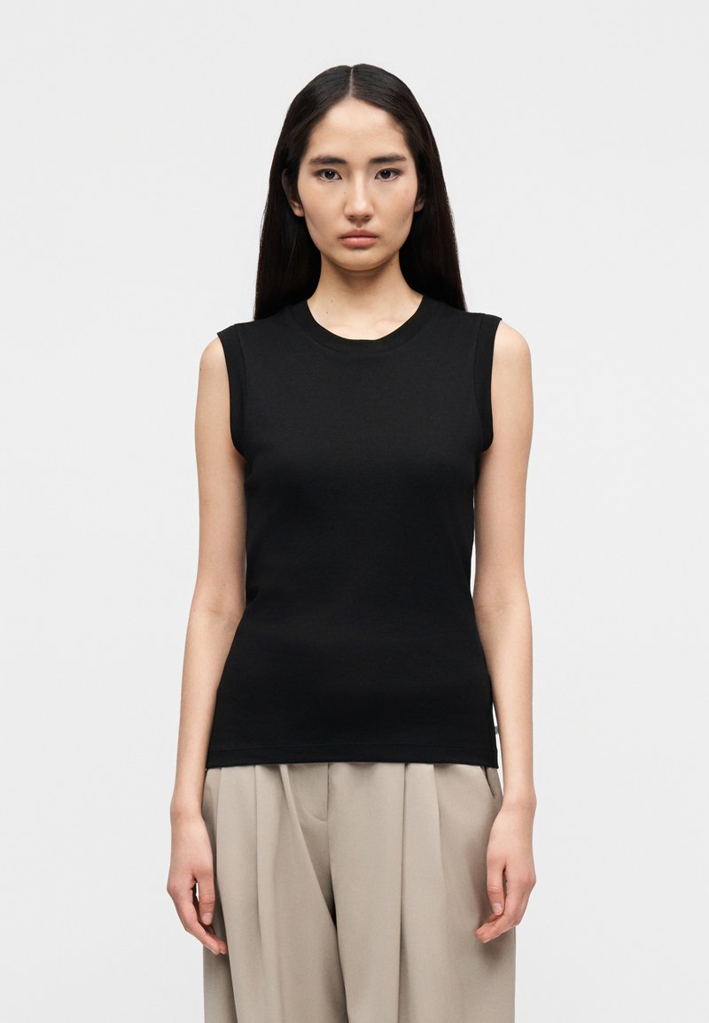 Γυναίκα με μακριά σκούρα μαλλιά πο υ φοράει μαύρο αμάνικο τοπ και μπεζ παντελόνι με φαρδιά πόδια, στέκεται μπροστά σε απλό λευκό φόντο.