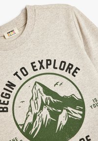 Hellgrauer Sweatshirt mit Rundhalsausschnitt, auf dem ein Bergmotiv in Dunkelgrün und der Text "BEGINNEN ZU ERKUNDEN" in fettem Schwarz zu sehen ist.
