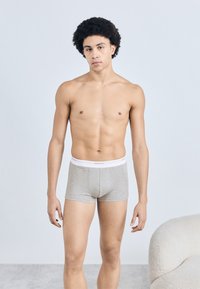 Jeune homme aux cheveux bouclés, debout pieds nus, portant un boxer gris dans une pièce minimaliste avec une chaise blanche texturée.