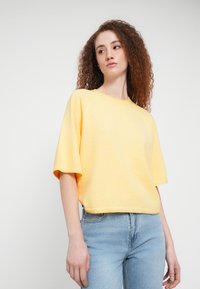 Κοντομάνικο, άνετης γραμμής κίτρινο τοπ από υφή υφάσματος, με στρογγυλή λαιμόκοψη και κοντό τελείωμα, συνδυασμένο με ανοιχτόχρωμο τζιν παντελόνι.