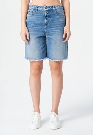 Shorts bermuda en denim bleu clair avec un ourlet effiloché. Présentent une taille haute, cinq poches et une fermeture à bouton. Stylés avec des baskets blanches.