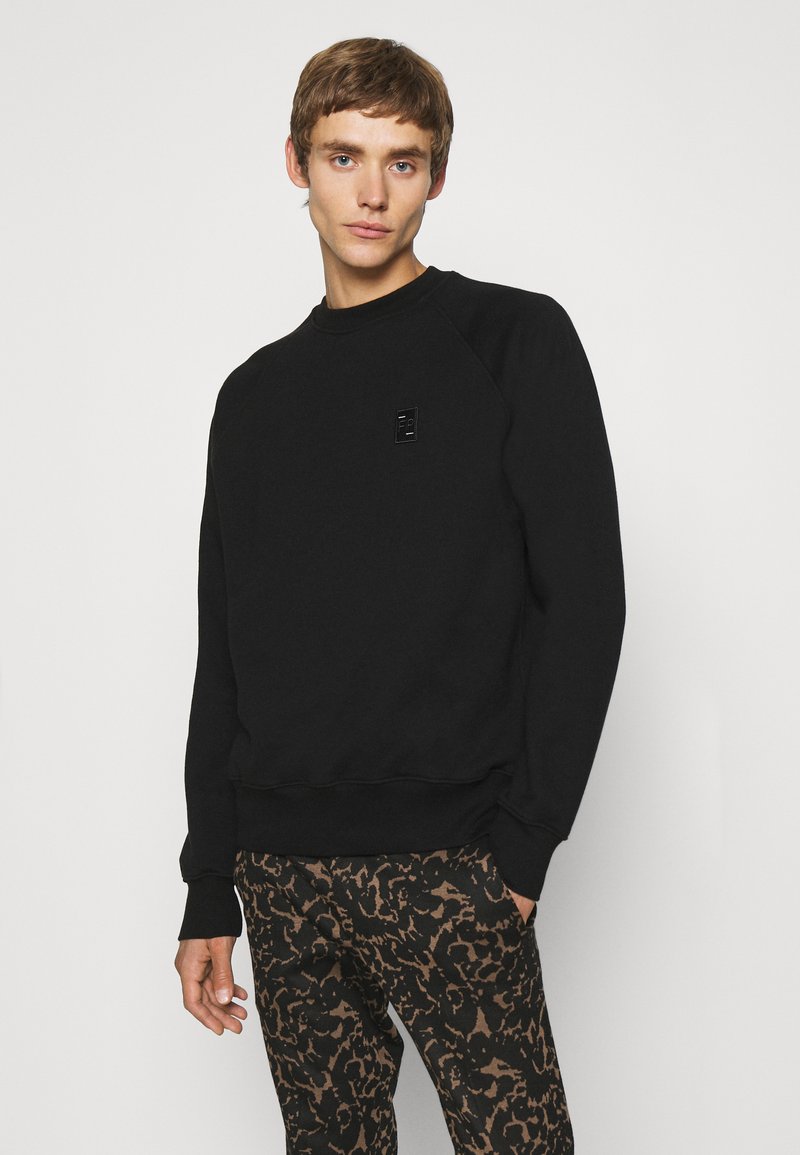 Sweatshirt noir avec un col rond et des manches longues, comportant un patch logo subtil. Assorti avec un pantalon à imprimé animal marron, mettant en valeur la texture et le design.