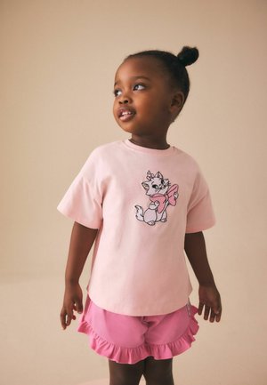 Fiatal lány rózsaszín pólóban, amelyen egy masnis rajzfilmmacska látható, és hozzáillő, fodros rózsaszín rövidnadrágban, oldalra néz egy semleges háttér előtt.