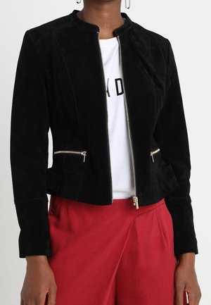 Chaqueta de ante negra con cuello redondo, diseño ajustado y bolsillos con cremallera. Llevada sobre una camiseta blanca y combinada con pantalones rojos.