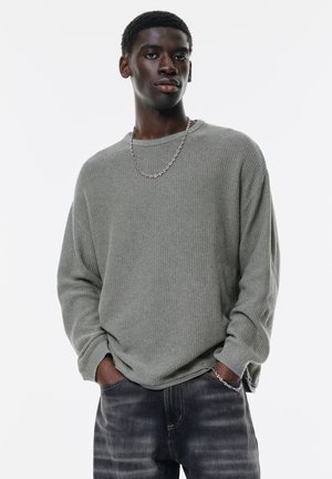 Gray waffle knit sweater with a relaxed fit, long sleeves, and a crew neckline, paired with dark denim jeans and a silver chain necklace.