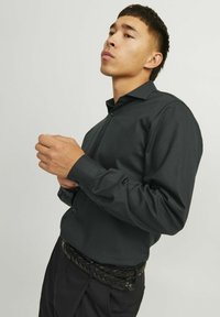 Camisa de manga larga gris oscuro con una textura sutil, cuello puntiagudo y puños con botones. Se lleva con un cinturón negro trenzado y pantalones oscuros a medida.