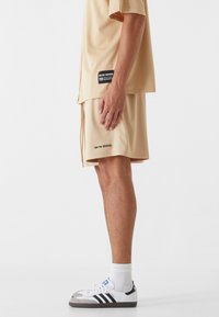 Ensemble de vêtements de sport en mesh beige, comprenant un haut à manches courtes et un short, avec des patchs de marque et un cordon de serrage. Porté avec des baskets blanches et des chaussettes.