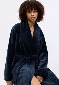 Tummansininen plush-kotitakki, jossa on saalahan, pitkät hihat ja sidottava vyötärö. Pehmeä tuntuma ja pitkä pituus, sopii rentoutumiseen tai yöpuvuksi.