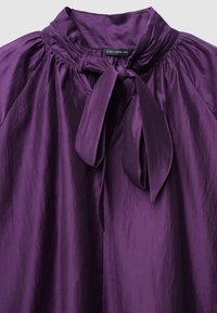 Blouse violette avec un col haut et un nœud, fabriquée en tissu lisse. Des plis le long des épaules ajoutent de la texture.