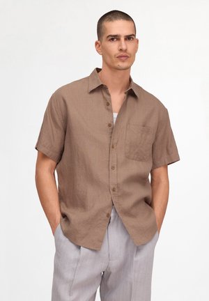 Mann mit kurzen Haaren, der ein braunes kurzärmeliges Hemd mit Knöpfen und hellgraue Hose trägt, steht mit den Händen in den Taschen vor weißem Hintergrund.