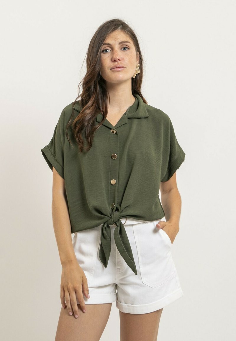 Blouse vert olive à manches courtes avec col et boutons sur le devant, dotée d'un détail de nœud à l'avant. Portée avec des shorts blancs à ourlet roulé.