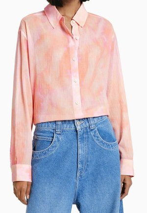 Woman wearing a pink and peach tie-dye long-sleeve button-up shirt paired with high-waisted blue denim jeans.