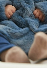Niebieski, puszysty, teksturowany koc częściowo przykrywa małe rączki i nóżki. Koc ma miękką, futrzaną powierzchnię bez widocznych wzorów.