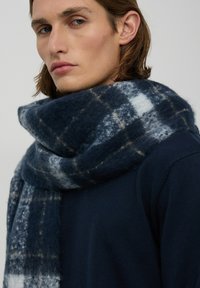 Granatowy szalik w kratkę wykonany z miękkiego materiału, z połączeniem ciemnoniebieskiego i jasnoniebieskiego oraz beżowymi akcentami, luźno owinięty wokół szyi.