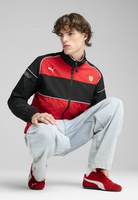 Giacca rossa e nera con zip, polsini elasticizzati e logo Ferrari; abbinata a jeans baggy azzurri chiari e sneaker rosse Puma.