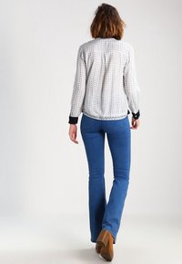 Camicetta bianca a fantasia con maniche lunghe e polsi neri, abbinata a jeans a zampa d'elefante blu e stivali con tacco quadrato color cammello.