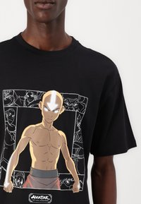Sort bomulds T-shirt med et grafisk print af en karakter med skaldet hoved, brun hud og grå øjne, sat mod en hvid baggrund.