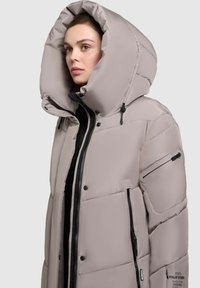 Lys grå pufferjakke med stor hætte, lynlås foran, sidelommer og sort hardware. Har kviltet struktur og logo detaljer.