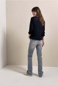 Mørkeblå bluse med opvalsede ærmer, parret med lysegrå jeans. Modellen står med ryggen til kameraet på en neutral baggrund.