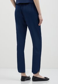 Personne portant un pantalon bleu marine fuselé avec une fente sur l'ourlet et des ballerines noires, se tenant sur un sol blanc devant un fond uni.