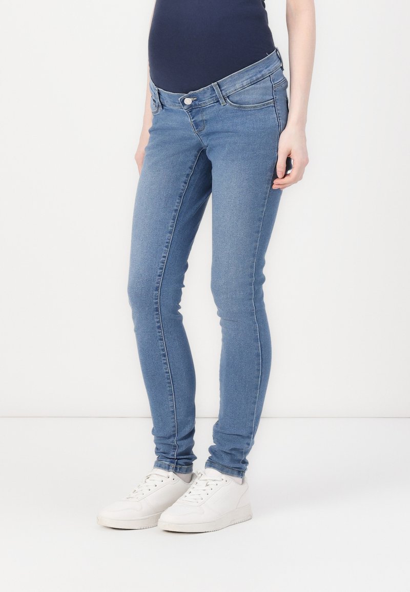 Skinny light blue jeans made of denim with a mid-rise waist, featuring a button closure and subtle pocket stitching. Paired with white shoes.